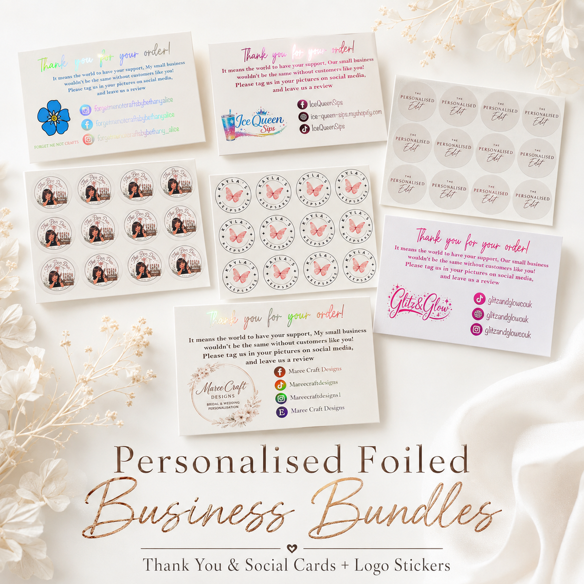 A flat-lay presentation of a personalized foiled business bundle, showing various printed cards, logo stickers, and round name tags with custom designs. The cards feature elegant gold, pink, and blue foil accents, and are arranged on a white fabric background decorated with dried white flowers.